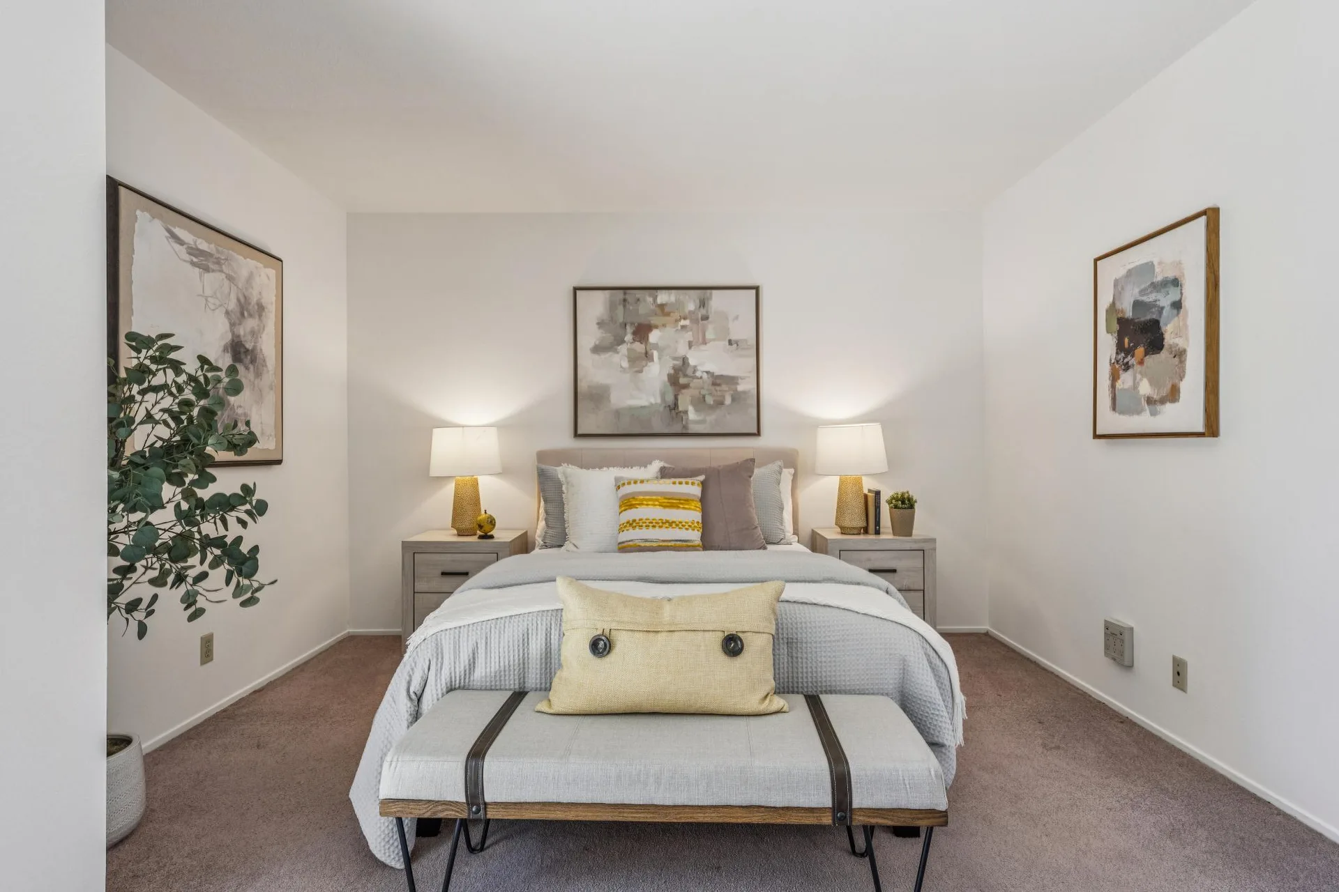 Second bedroom with neutral tones, warm lamp lighting, bench at foot of bed, and plush carpet