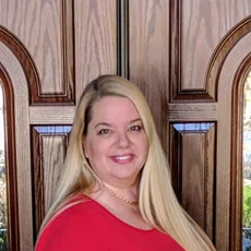 Monica Franks, REALTOR®, professional portrait in a red top, standing in front of arched wooden doors