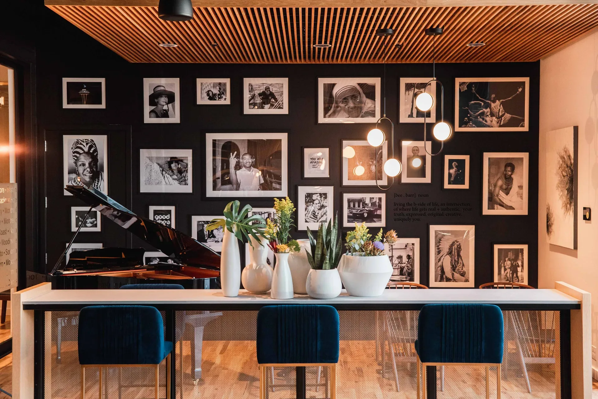 Interior gallery lounge at Realogics Sotheby's International Realty Bellevue — matte black gallery wall, teal bar stools, grand piano, wood slat ceiling with chandelier