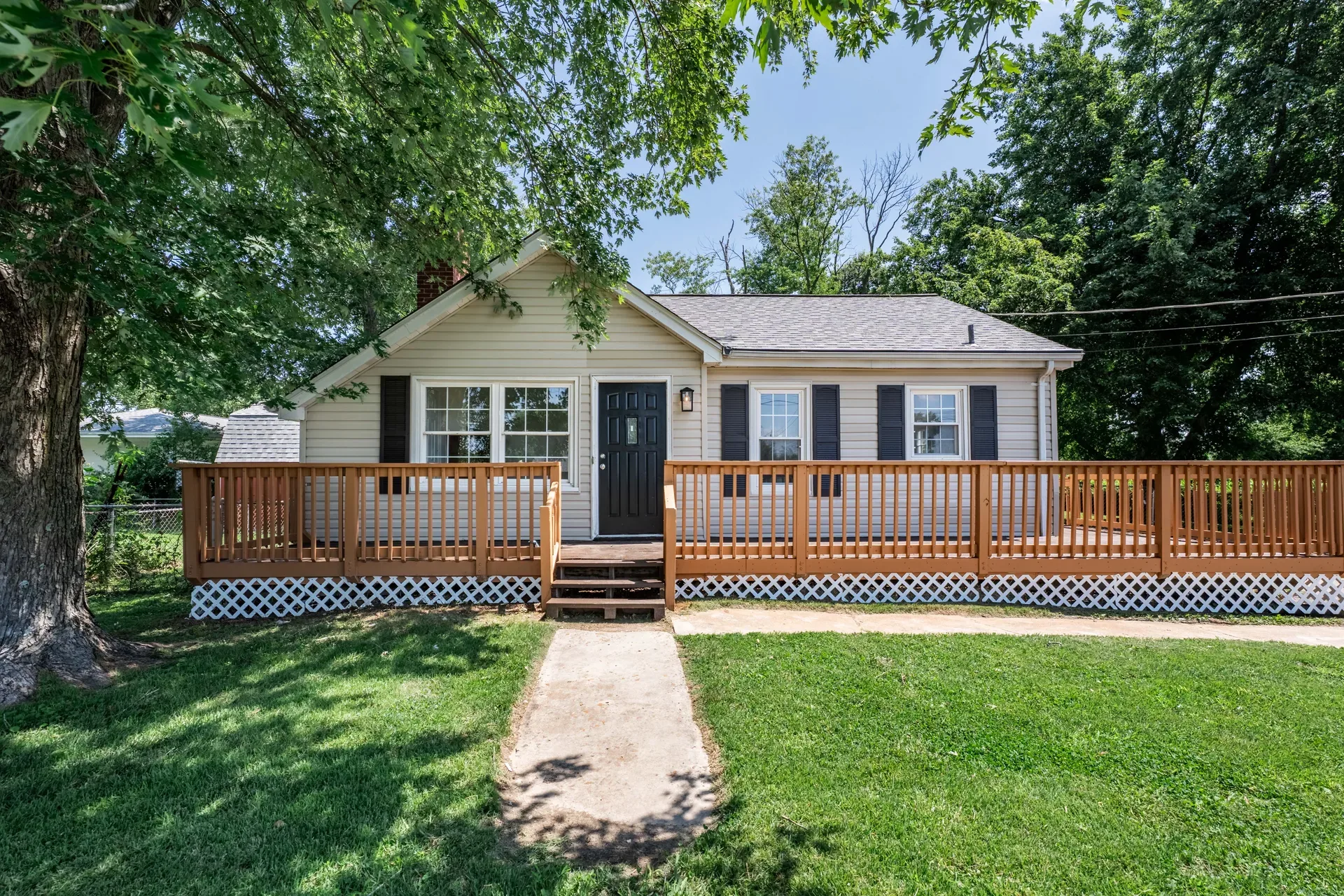 Front exterior of the ranch home with large wooden wraparound deck, beige siding, and dark shutters