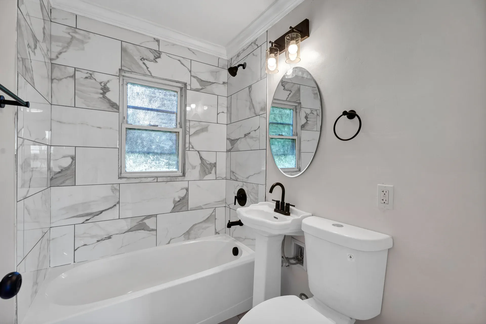 Renovated main floor bathroom with large format marble-look wall tiles, pedestal sink, oval mirror, and matte black fixtures