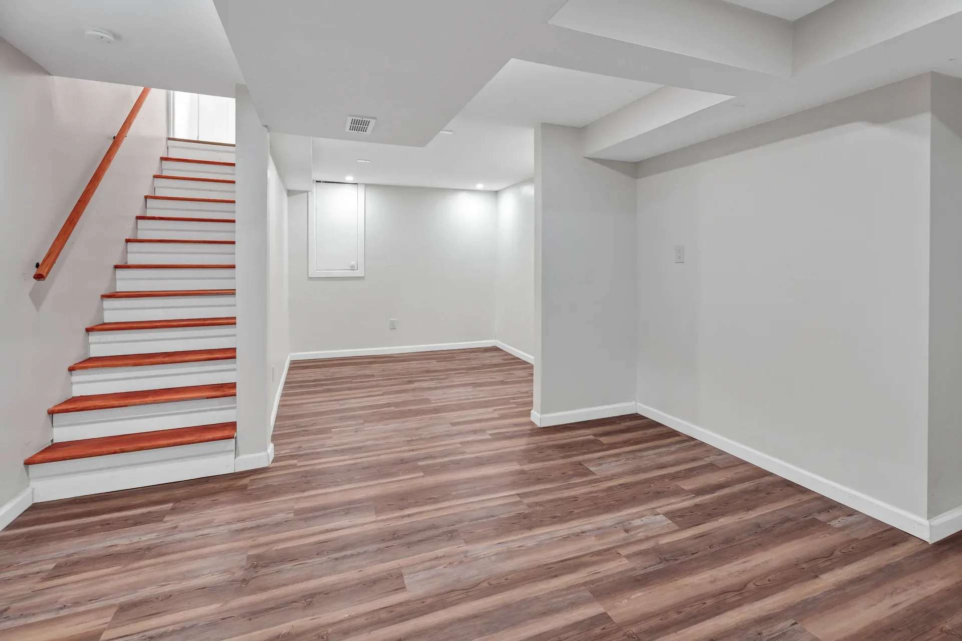 Finished basement recreation area with light gray walls, dark wood-look LVP flooring, stairwell, and recessed lighting