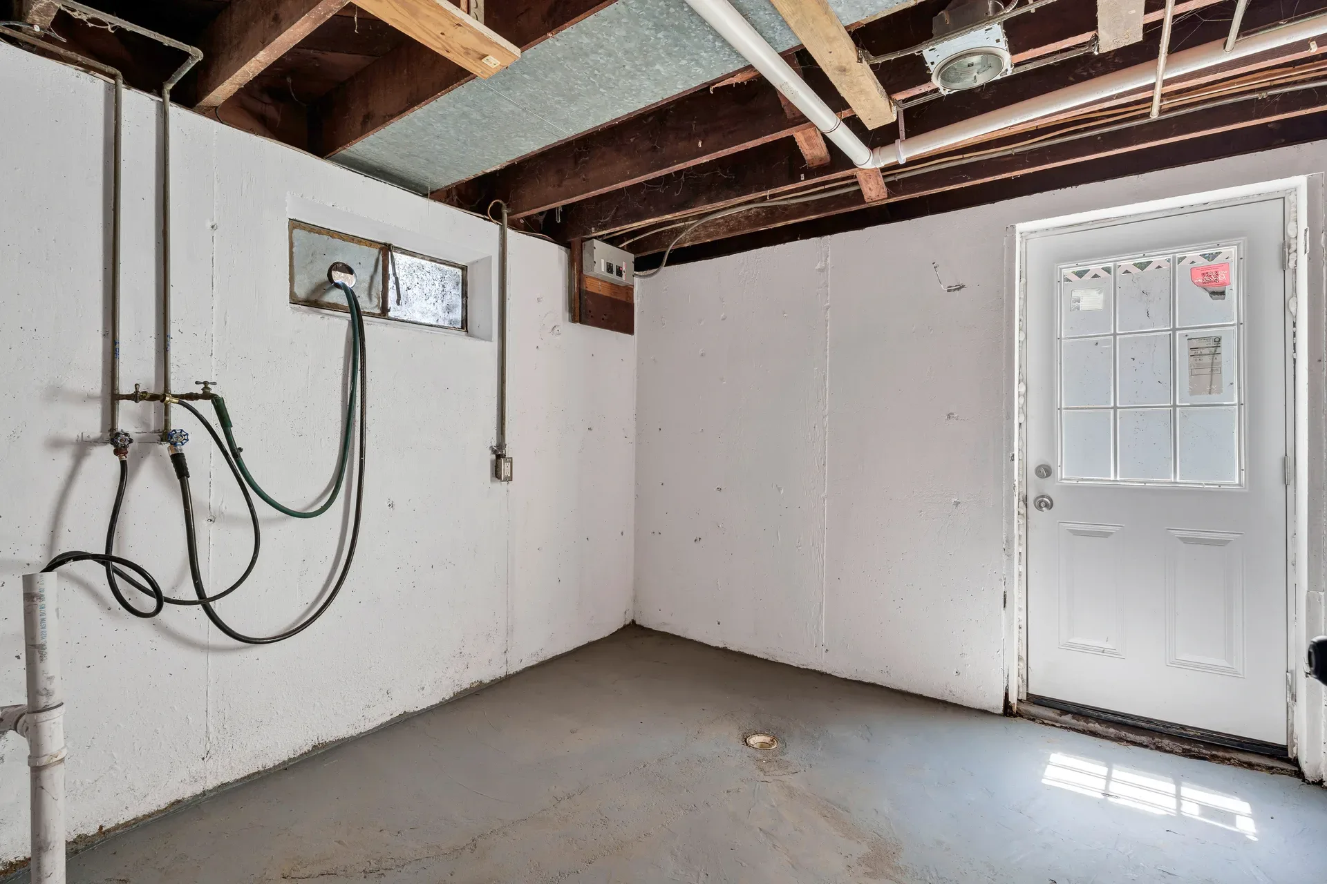 Walk-up basement utility area with painted concrete walls, exposed ceiling joists, daylight through exterior door