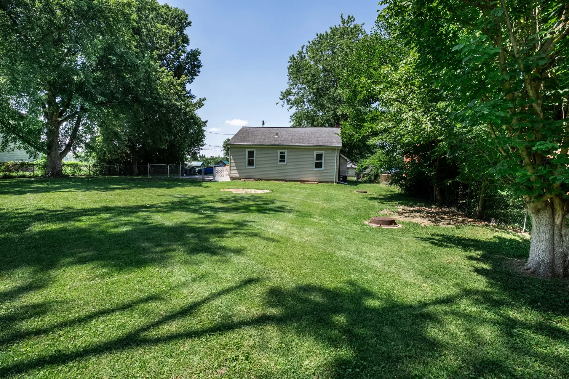 Spacious backyard view showing rear of the ranch home, large maintained lawn, mature trees, and chain-link fence