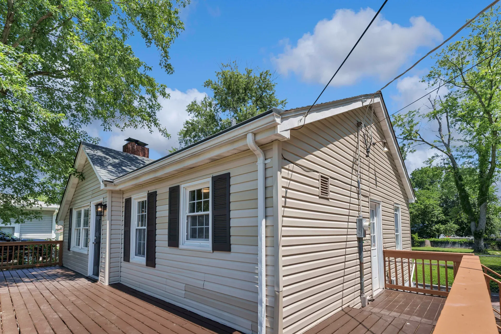 Side deck view of the ranch home with beige siding, brown wooden deck, white trimmed windows with dark shutters