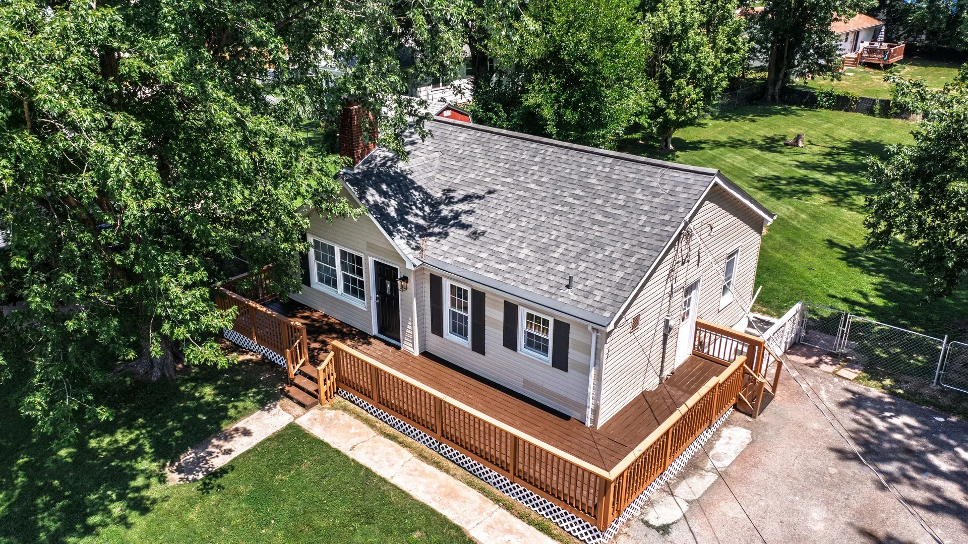 Aerial bird's-eye view of the ranch home showing yard, large wooden deck, and mature trees surrounding the property
