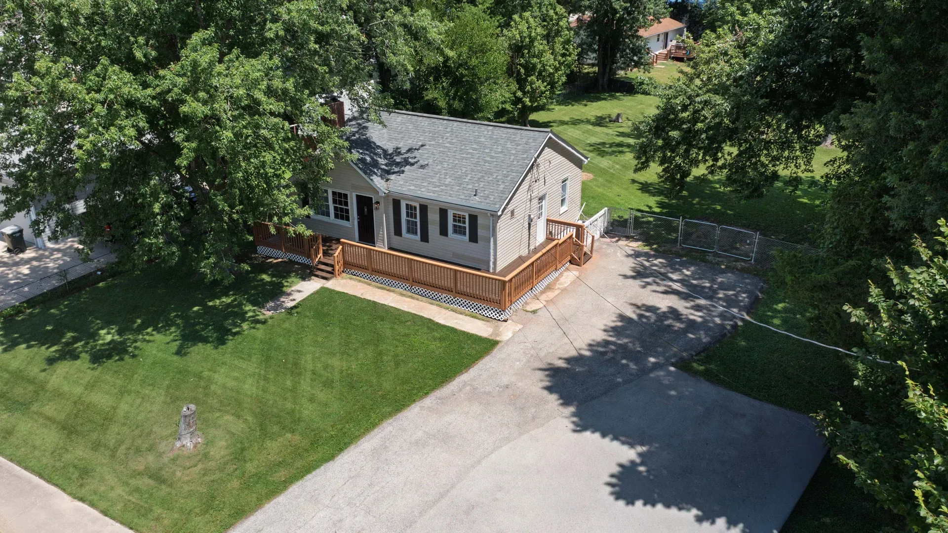 Aerial view of the single-story ranch home showing vinyl siding, shingled roof, wraparound deck, and tree canopy
