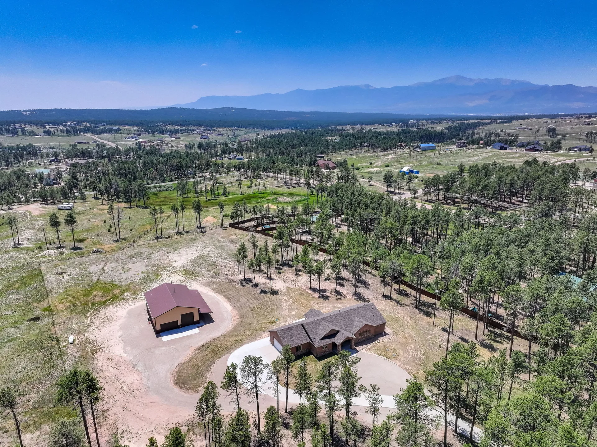 Aerial view of 6905 Wildridge Rd., a ranch home and workshop set in a mountainous, forested landscape in Black Forest, Colorado Springs, under a bright blue sky