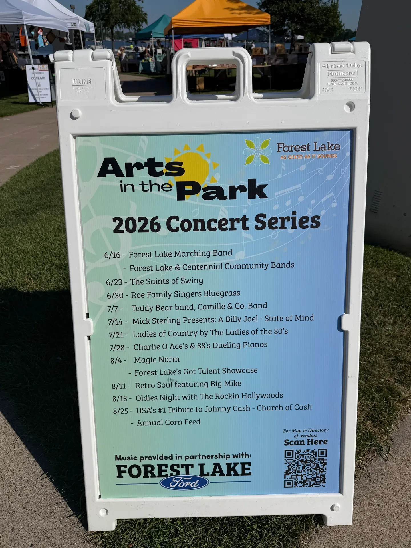Forest Lake Arts in the Park 2026 Concert Series sign listing the full schedule from June 16 to August 25, with acts including the Forest Lake Marching Band, Saints of Swing, and Roe Family Singers Bluegrass