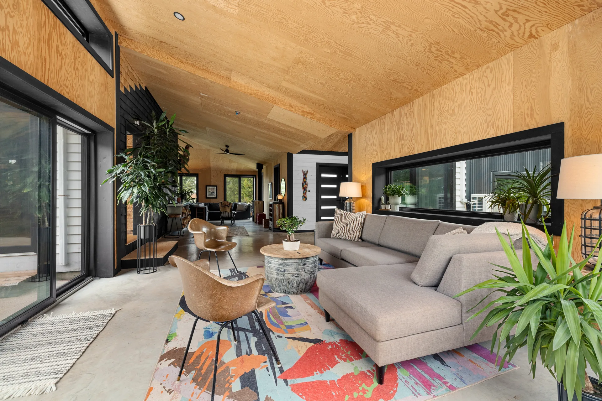 Modern living room addition with plywood-paneled walls, oversized black-framed windows, and concrete flooring