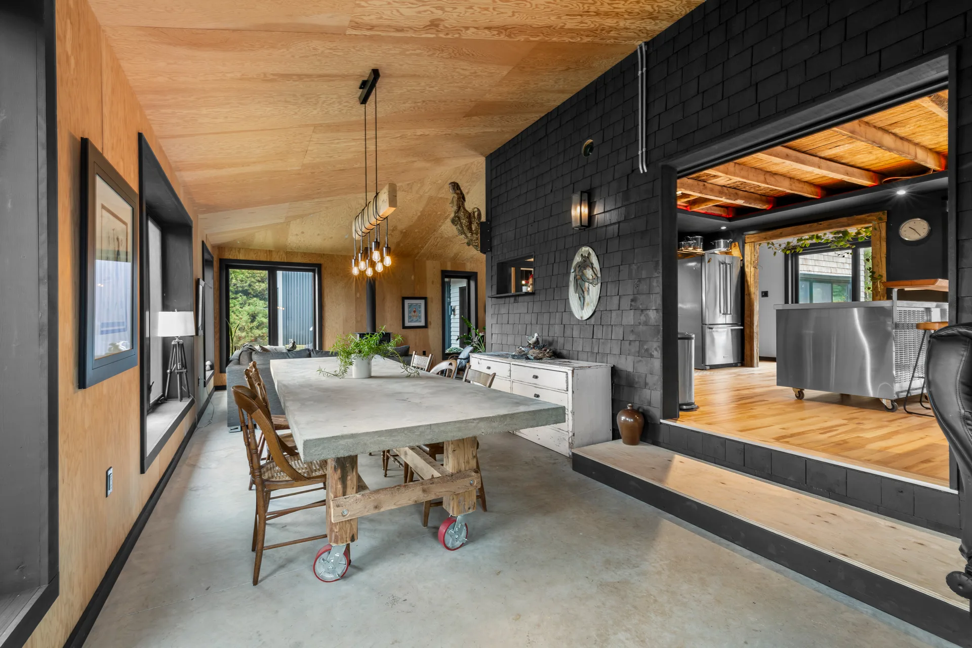 Dining room area in the barn-inspired addition with plywood-paneled walls, black-shingled accent wall, and concrete floor