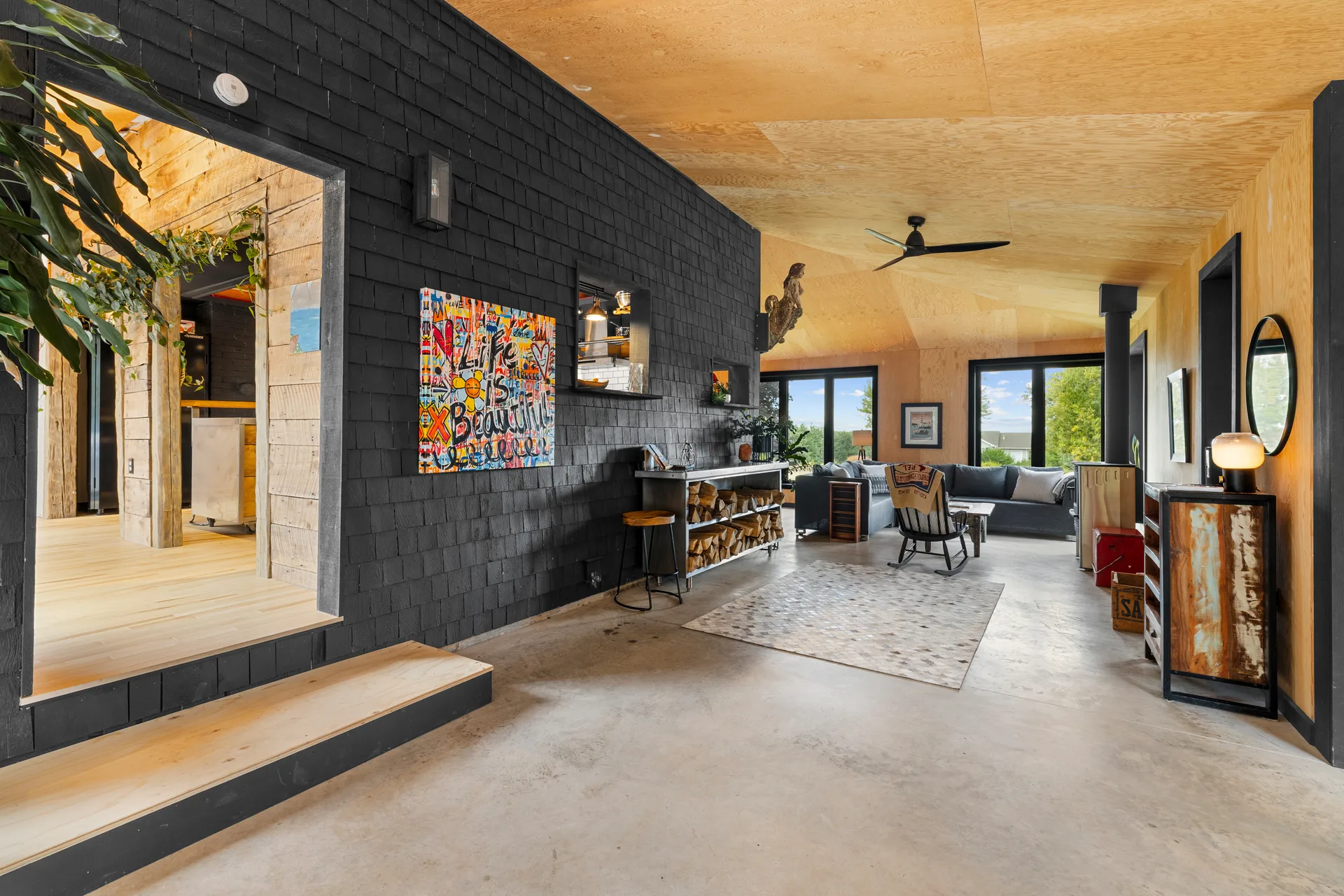Open-concept living room with exposed plywood ceiling, concrete floors, and black shingle feature wall