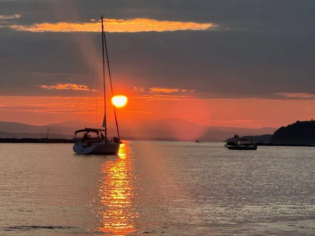 The sun dips below the horizon as a sailboat glides across Burlington Harbor