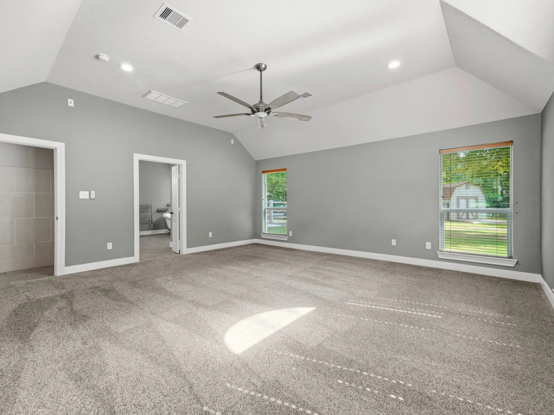 Master bedroom with vaulted ceiling, grey walls, carpeted flooring, and two windows with natural light