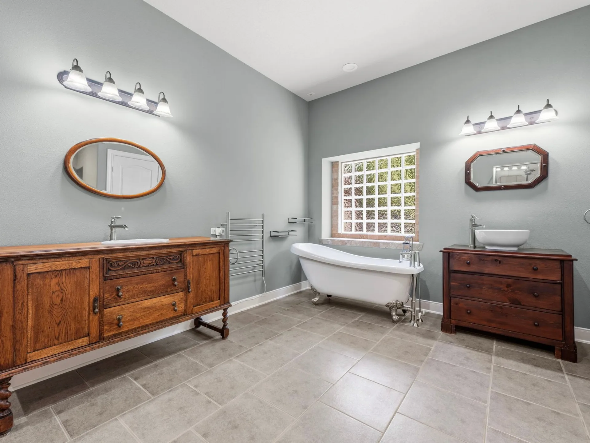 Master bathroom with two wooden vanities, clawfoot tub, gray tile flooring, and grid-style window