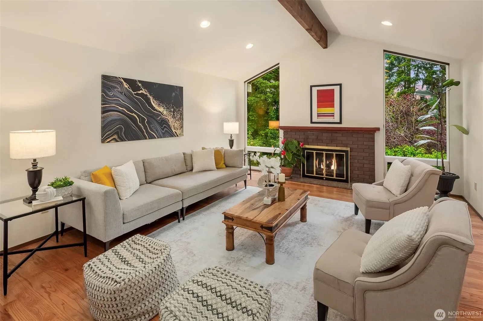 Vaulted living room with exposed wood beam ceiling, red brick fireplace, and hardwood floors flowing into the kitchen