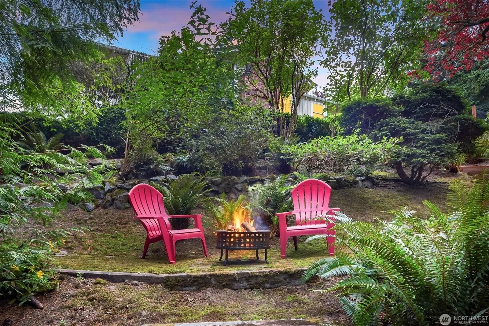 Backyard at dusk with a glowing fire pit, two red Adirondack chairs, and mature trees creating a cozy outdoor retreat