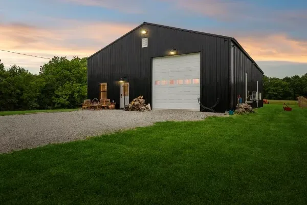 Modern metal barn facade with dark vertical siding, white garage door, and gravel driveway at twilight