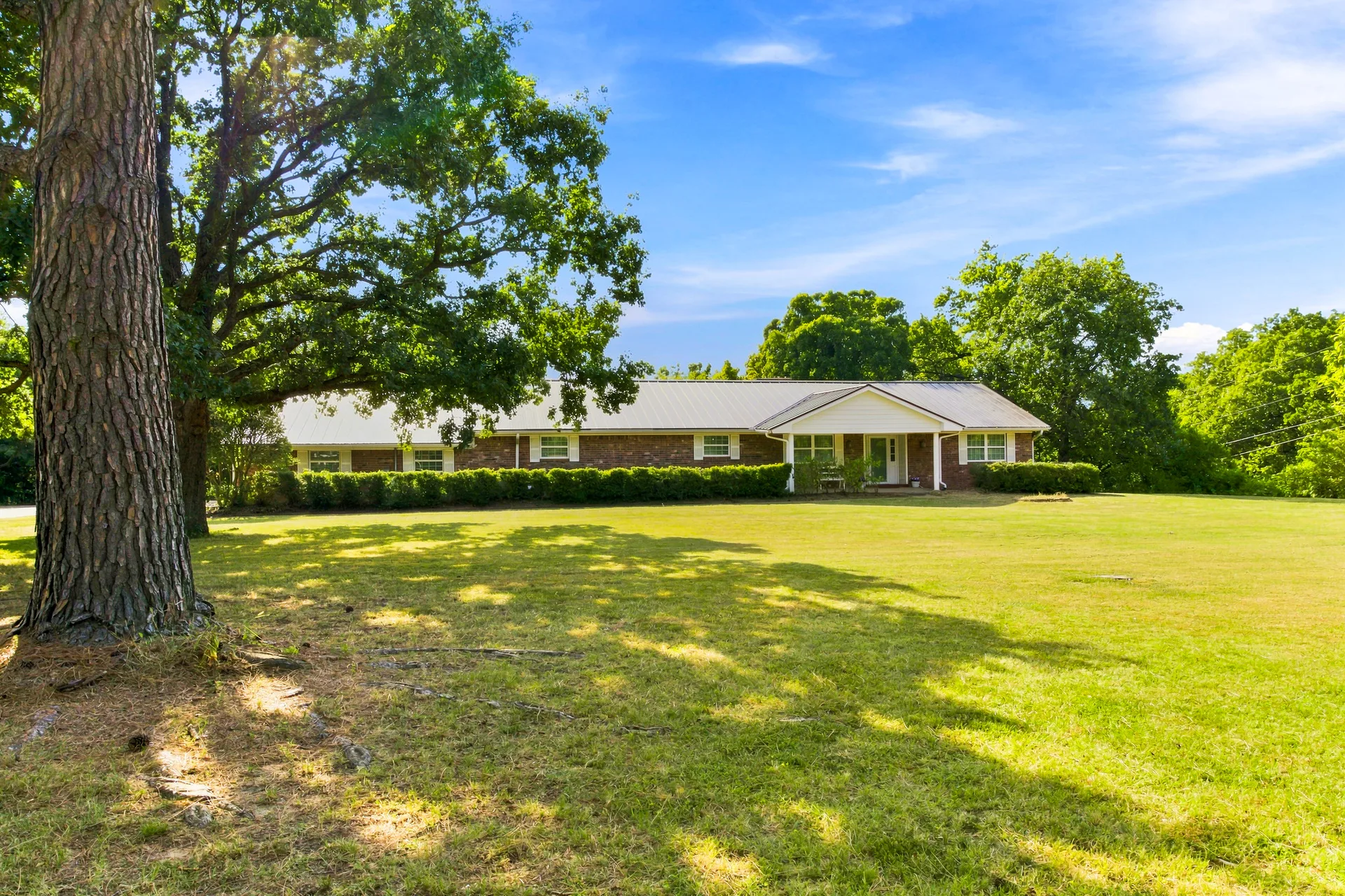 Front facade of 4723 Theresa Dr framed by mature oaks and pines, with a gabled porch entry, white trim, and long shadows cast across the lawn by the tree canopy