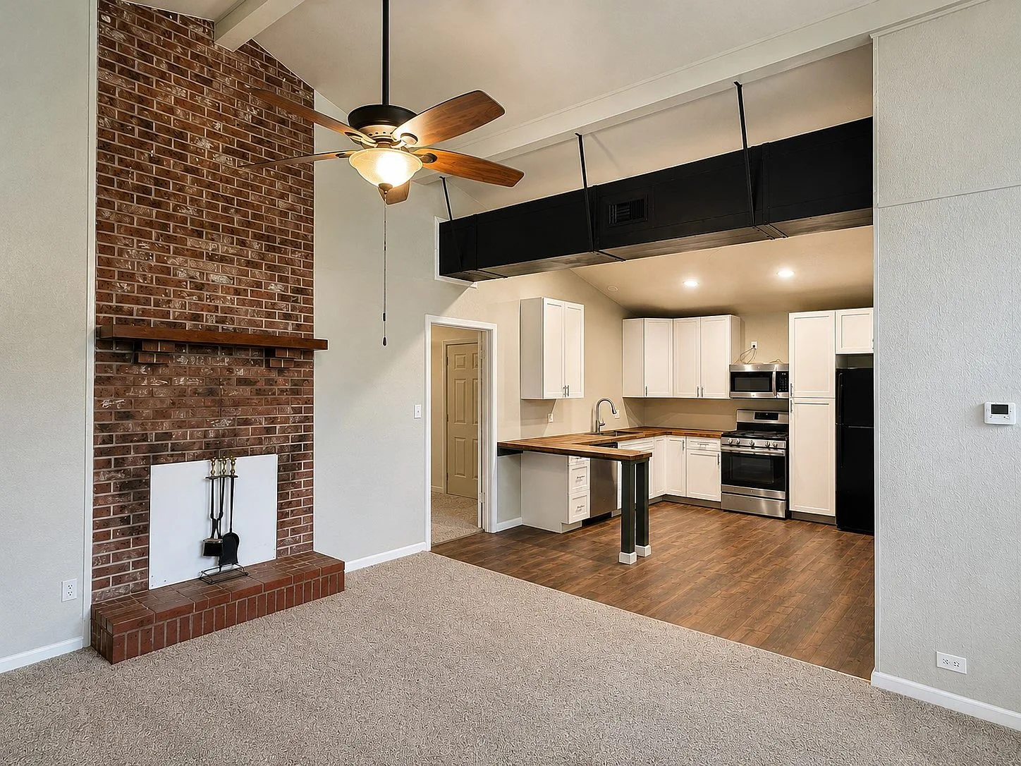 Open living room with vaulted ceilings and brick fireplace flowing into the kitchen
