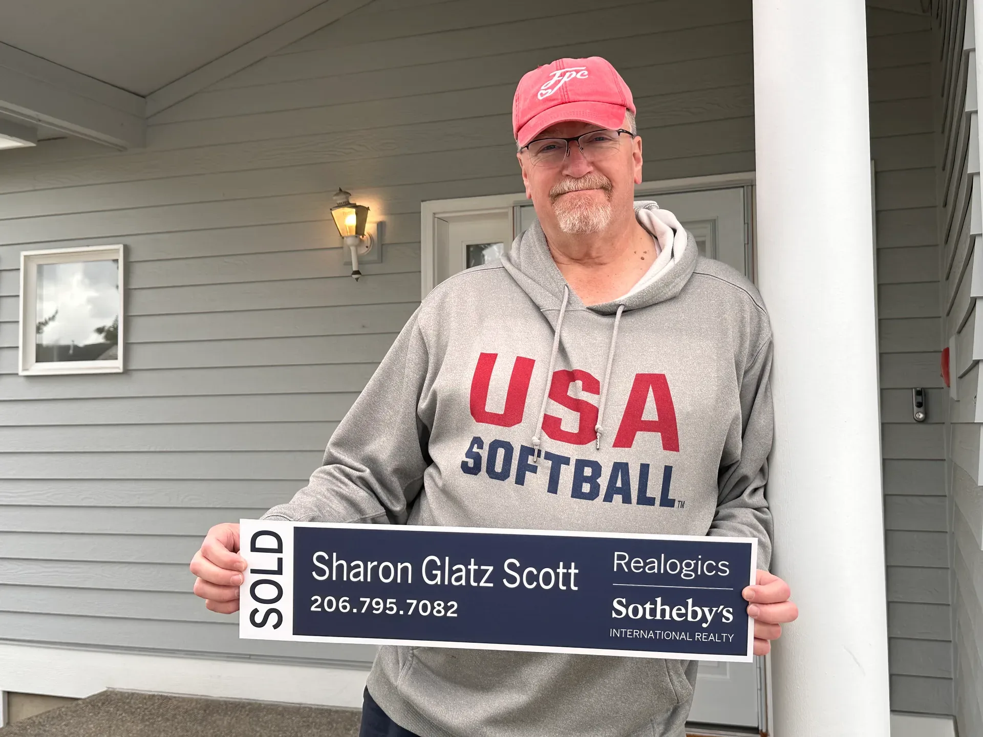 George holds a SOLD sign on the front porch of his new Eastside home, a light grey house with warm afternoon light