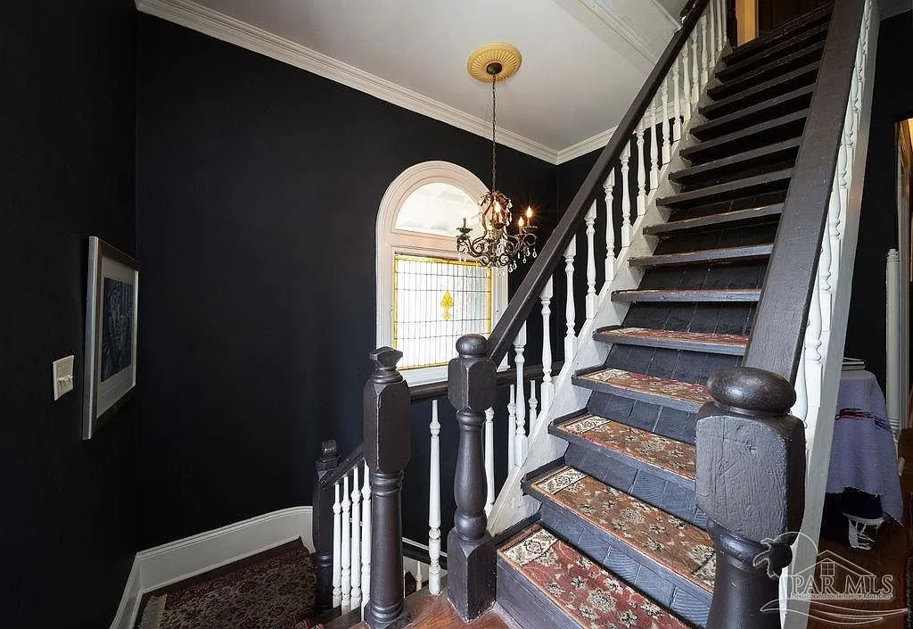 The grand staircase with white balusters and rug runners, lit by a stained glass window with an arched transom.