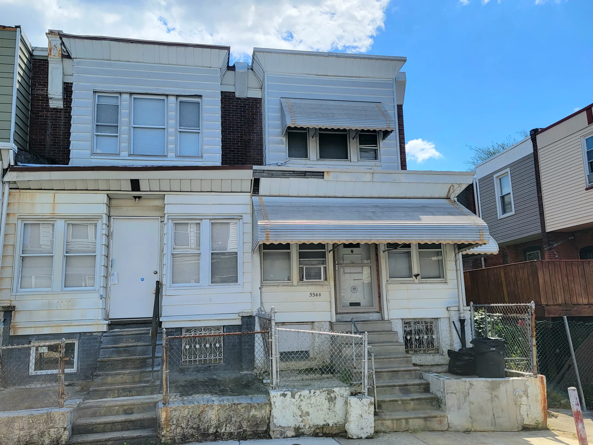 Two attached row homes in Northeast Philadelphia with aluminum siding and stone foundations under a bright blue sky