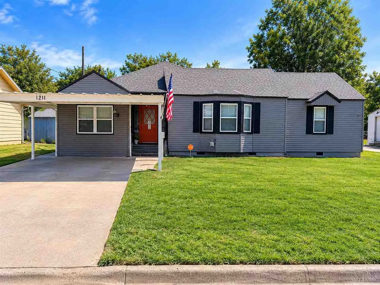 Front exterior of 1211 NW Parkview Blvd, a single-story gray ranch home with a bright coral front door and clear blue sky