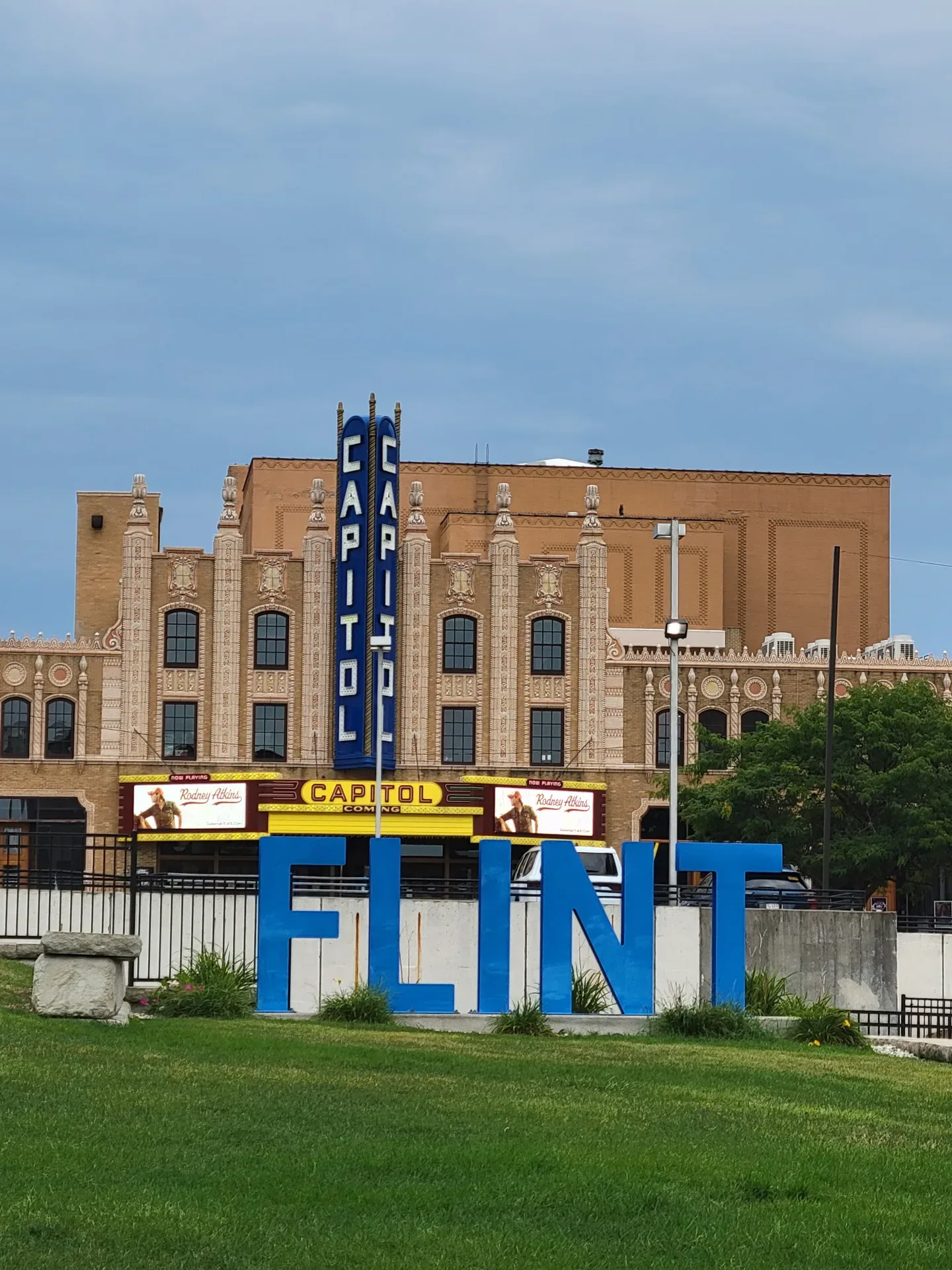 Exterior landmark view of the FIM Capitol Theatre in downtown Flint, with its historic brick facade, large blue FLINT lettering, and towering vertical sign beneath a cloudy sky