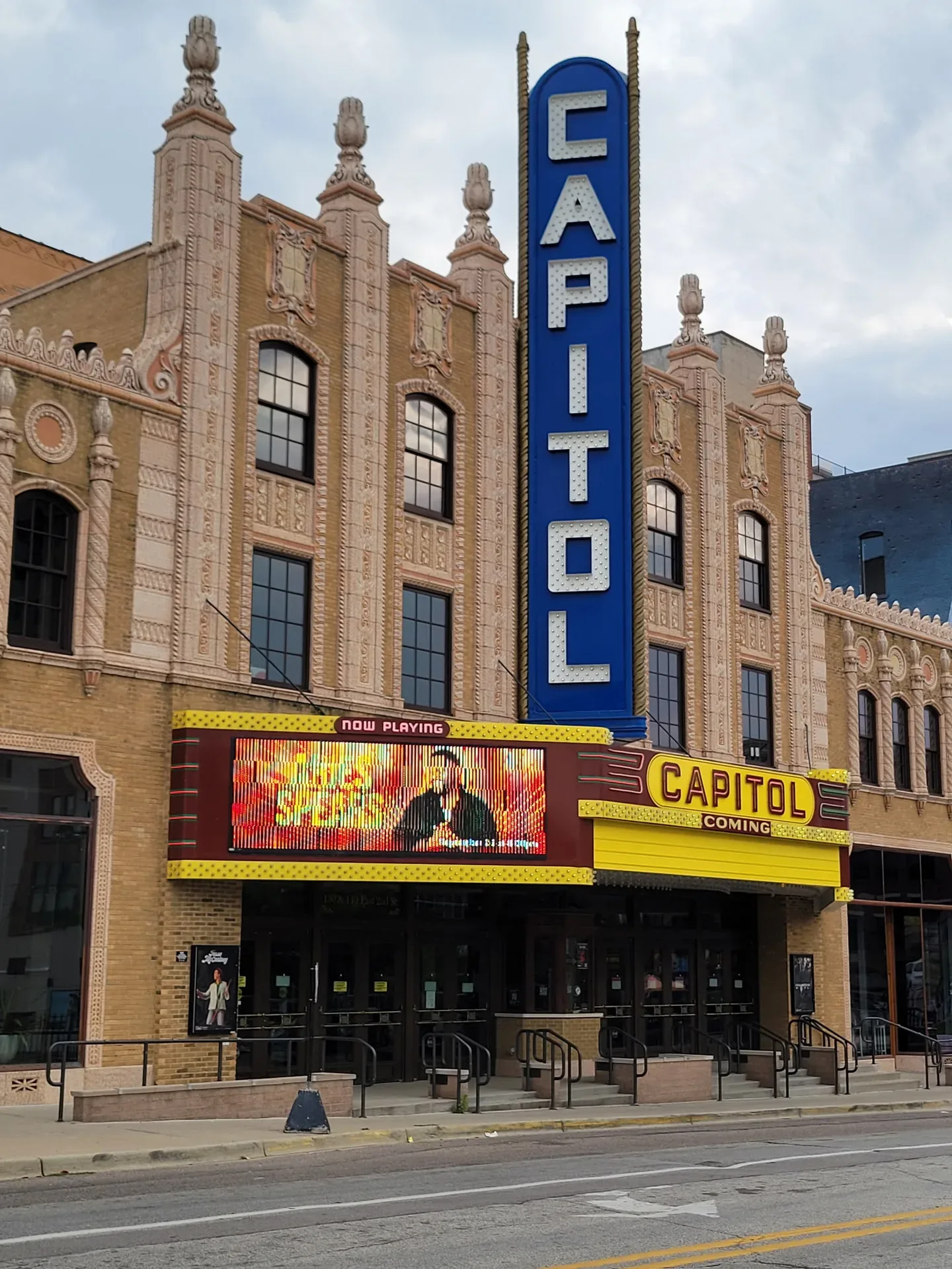 The historic street-level facade of the FIM Capitol Theatre in downtown Flint, with its digital marquee, vertical blue neon-style sign, glass storefront doors, and classical brick and masonry detailing under a cloudy sky