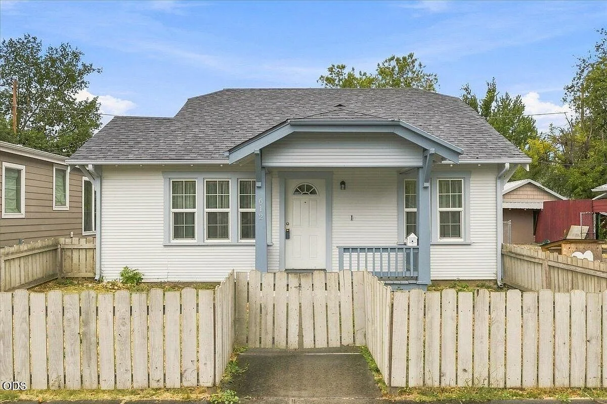 Front facade of 612 S Ivy St, a white bungalow with light blue trim, a covered porch, and a white picket fence