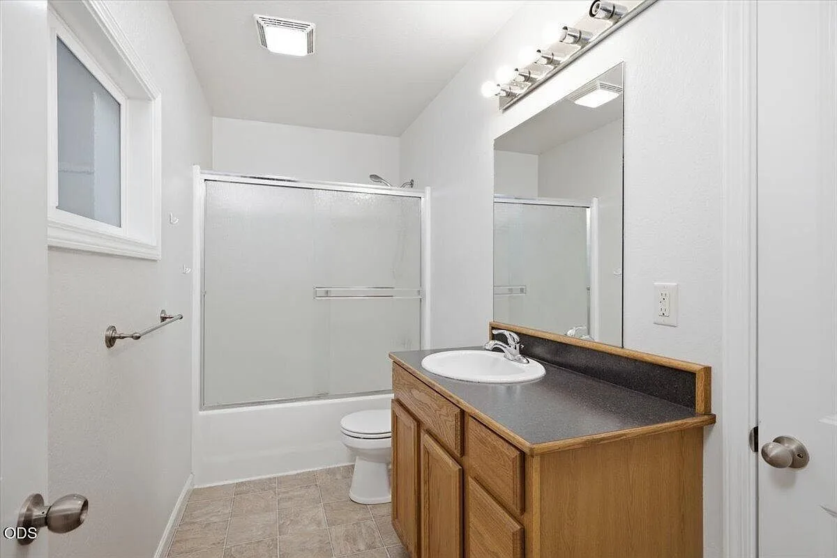 Full bathroom with oak vanity, dark countertop, and shower tub combo with glass doors