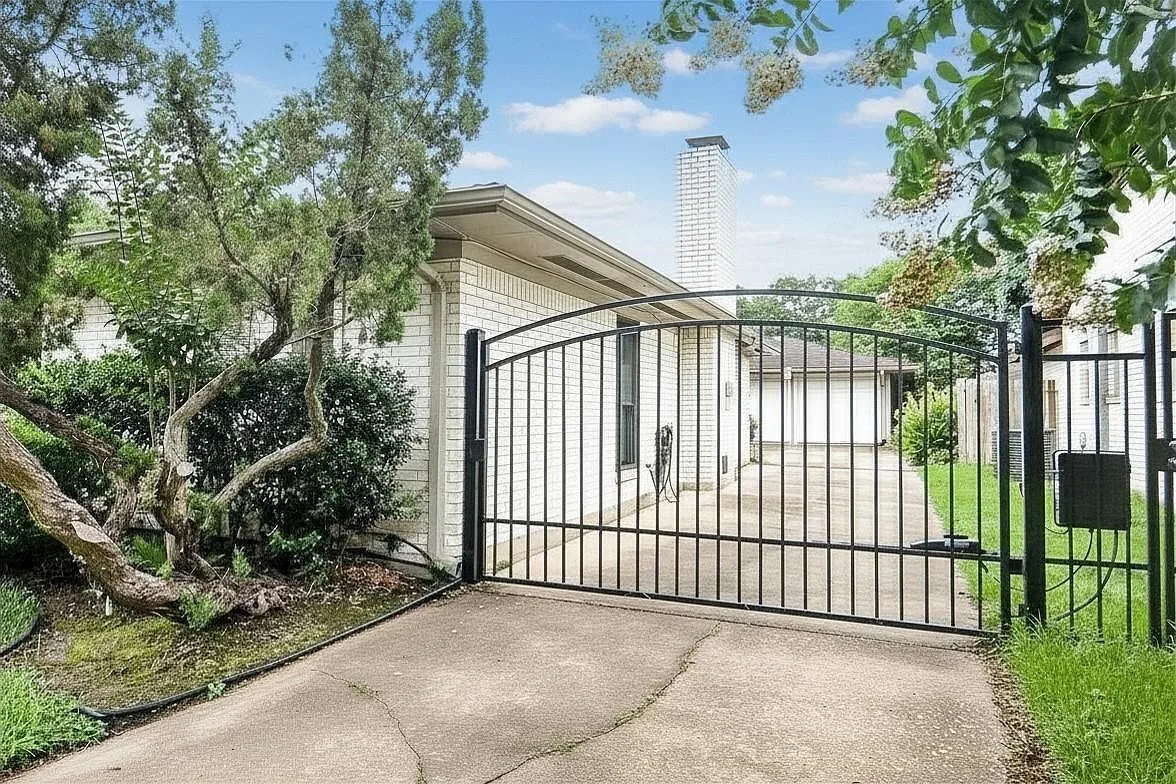 Side view of the gated driveway at 6203 Wigton Dr with a tall brick chimney and mature landscaping