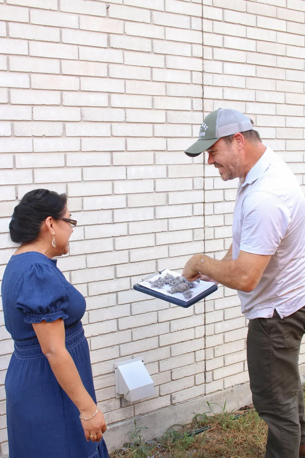 A home inspector kneels beside a vent on the white-painted brick exterior wall of a San Antonio home while his client watches, the kind of perimeter walk-around that happens during a wood-destroying insect inspection
