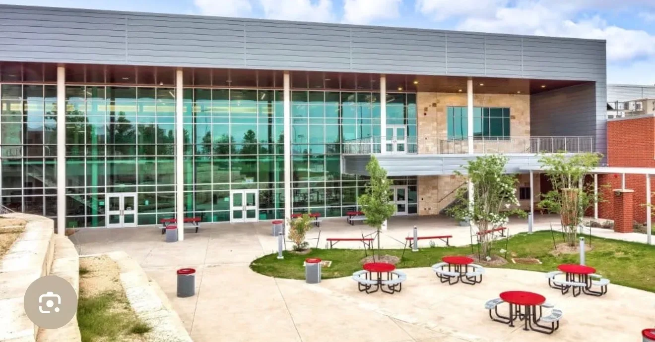 A modern school campus building with glass curtain walls, light stone accents, and a paved courtyard with young trees under a blue sky