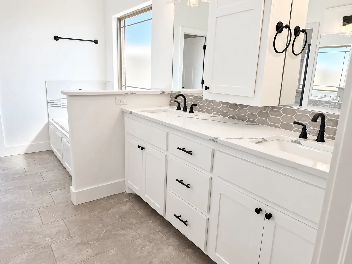 Bathroom with dual white vanities, marble-look countertop, hexagon tile backsplash, and matte black hardware