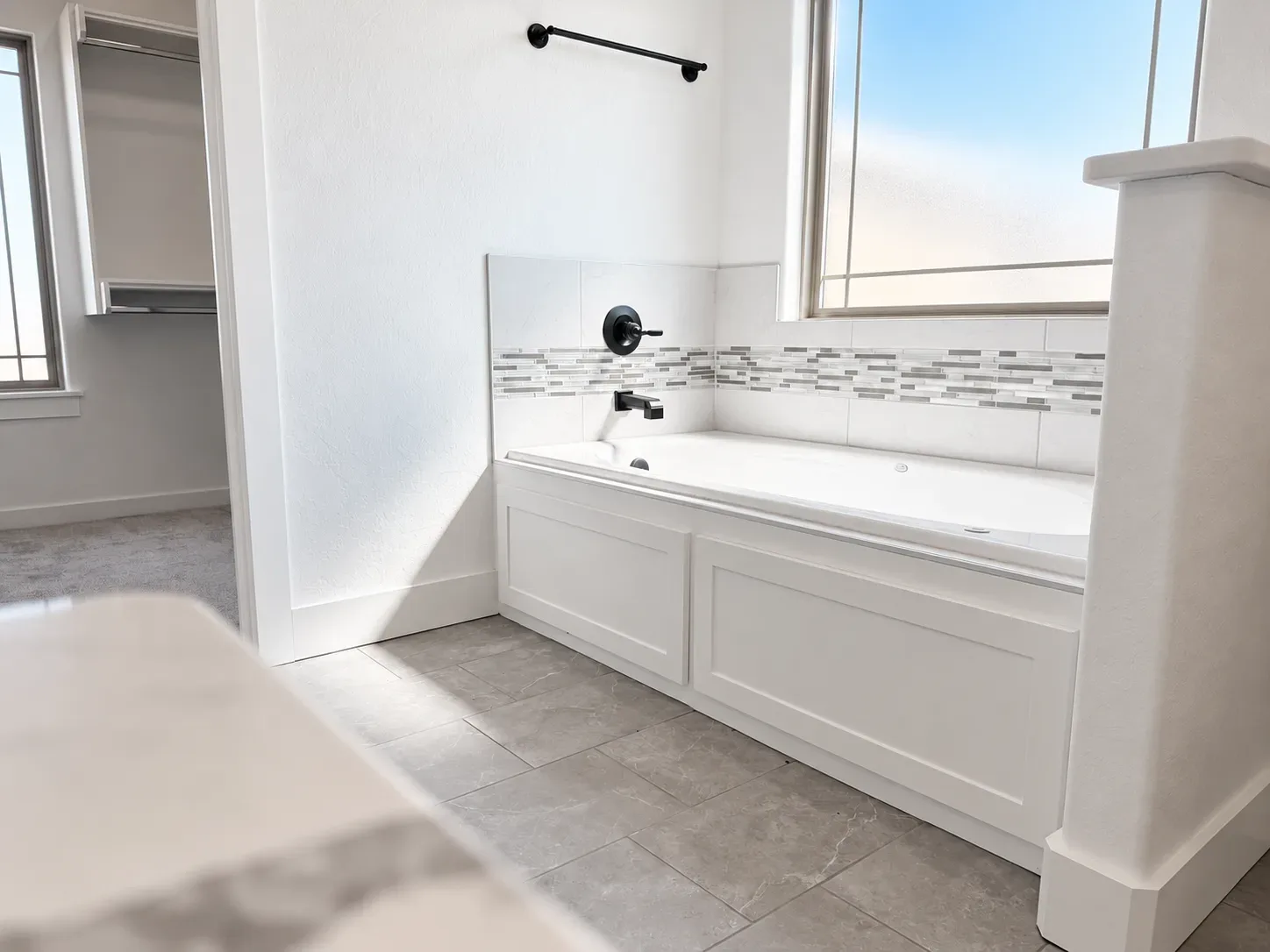 Bathroom with white soaking tub, matte black fixtures, gray tile flooring, and mosaic tile accent border