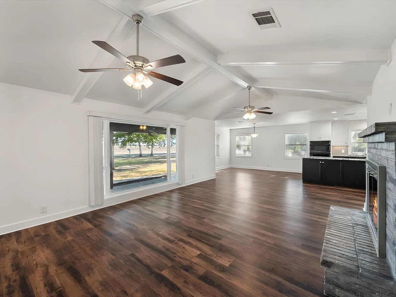 Open living area flowing toward the kitchen with exposed beams and gray brick fireplace
