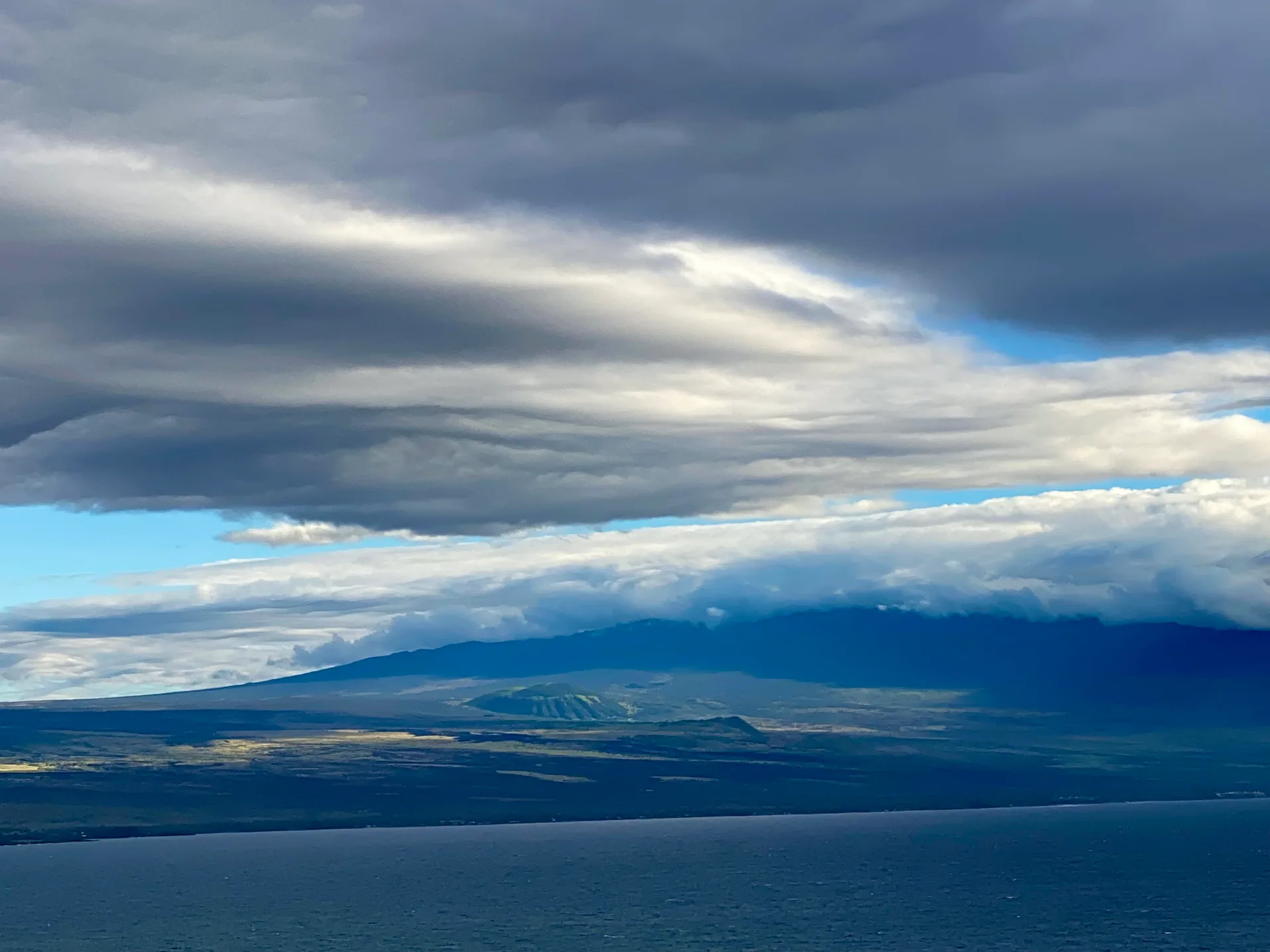 Volcanic mountain slopes meeting the ocean under cloud cover, with patches of sunlight on the land, on Hawaiʻi Island