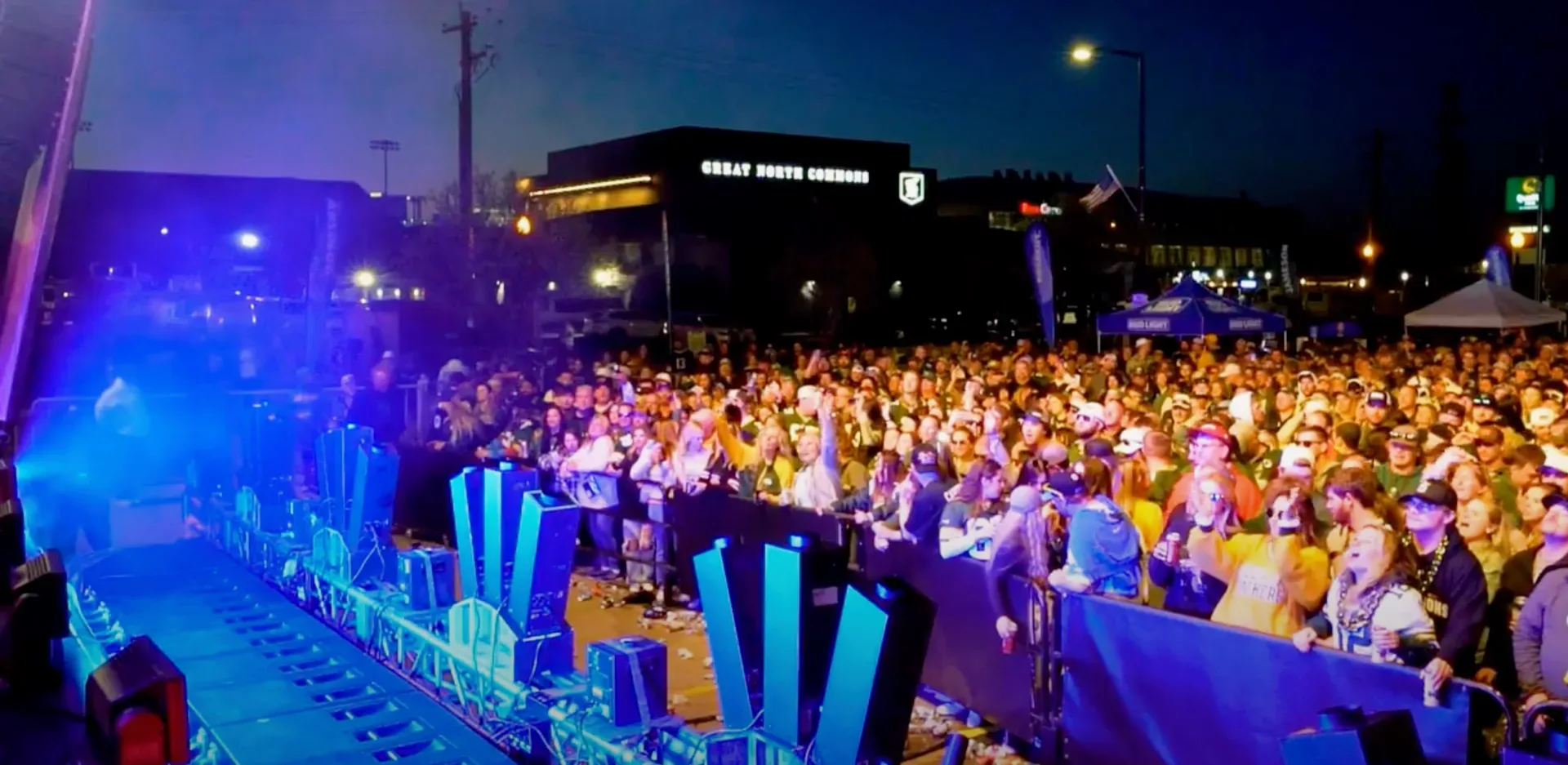 A large crowd at an outdoor concert in Bowling Green at night, the stage lit in blue behind the barriers