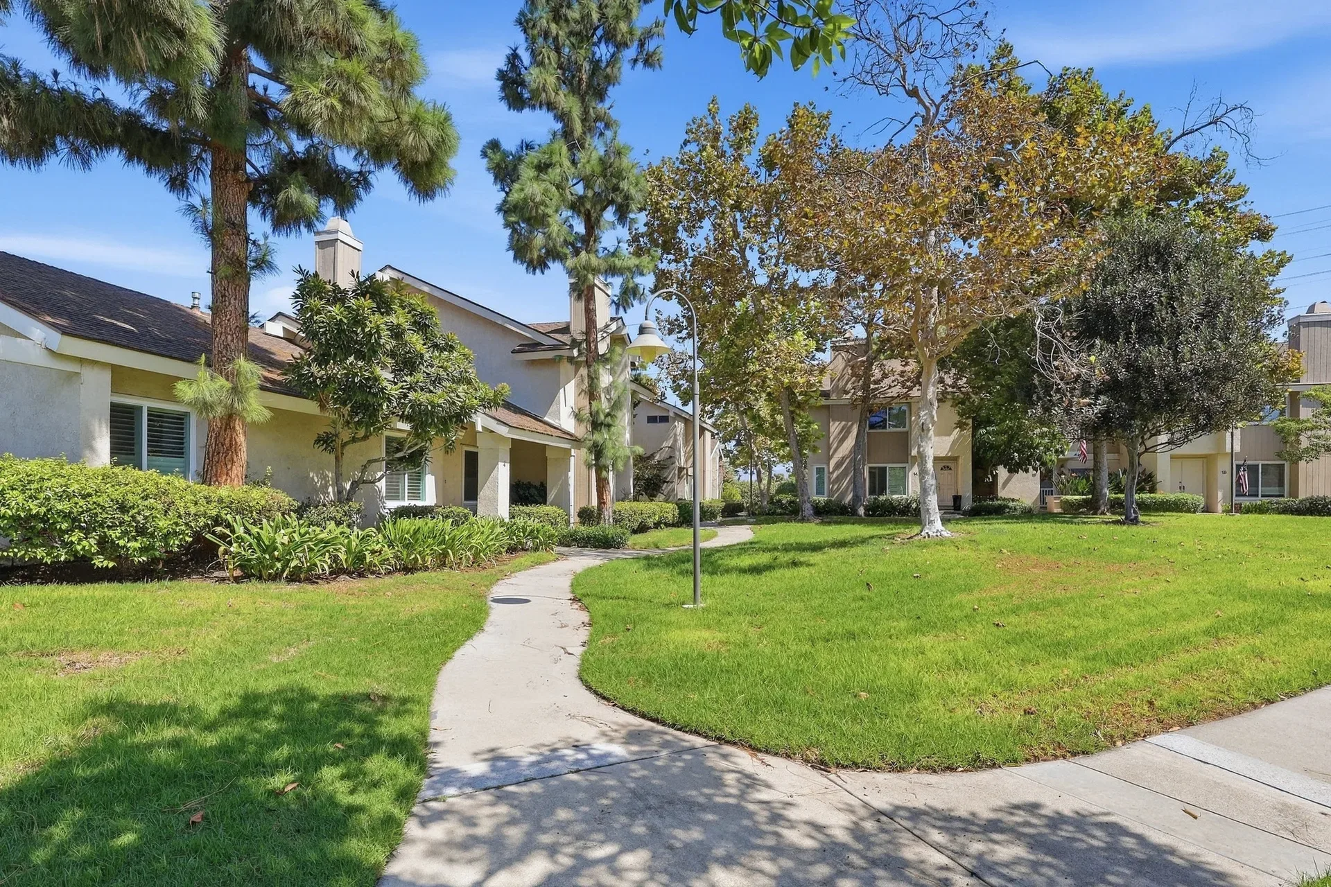 Community walkway and grounds around 8 Silverwood with stucco and wood-siding homes, mature landscaping, and a clear sky in Irvine