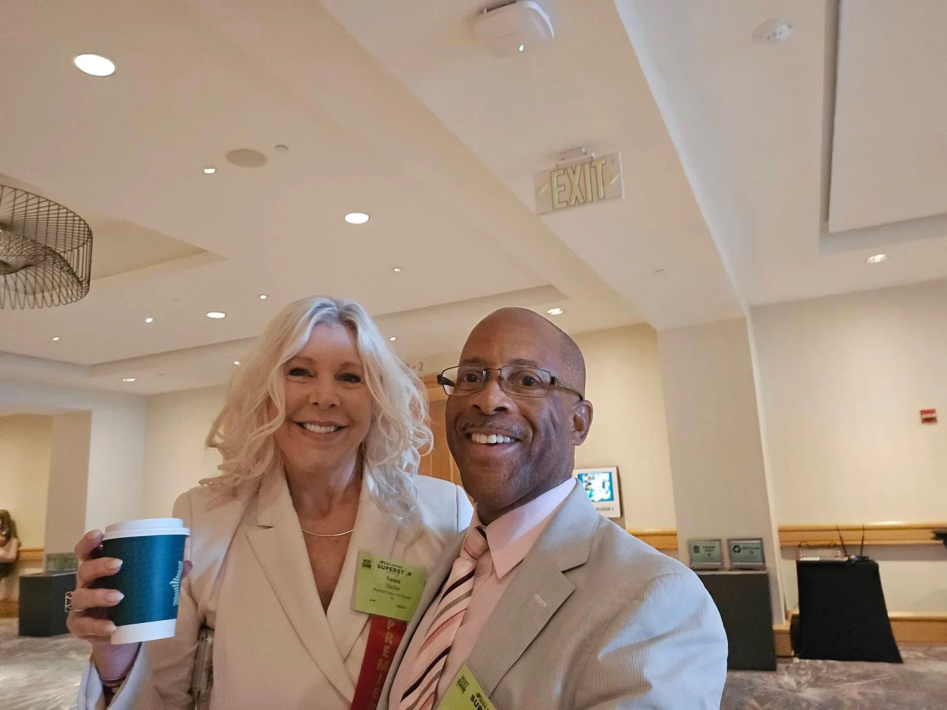 Robert Clarke takes a smiling selfie with a fellow conference attendee, a woman with blonde hair in a light blazer holding a green coffee cup, both wearing lime-green conference name tags at the Mike Ferry 2026 Superstar Retreat