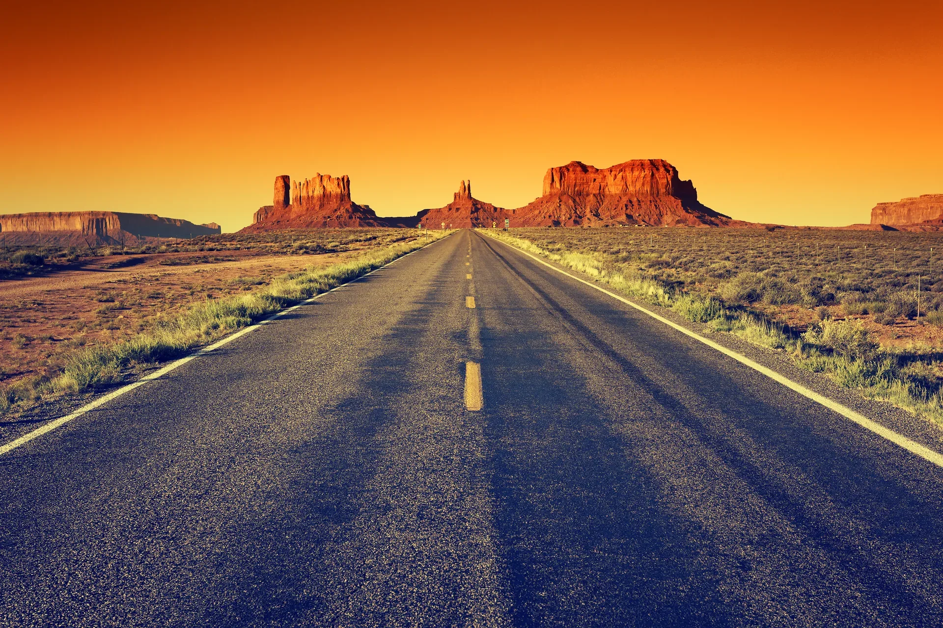 A long desert road running toward red rock mesas under a vivid orange sunset sky in northern Utah
