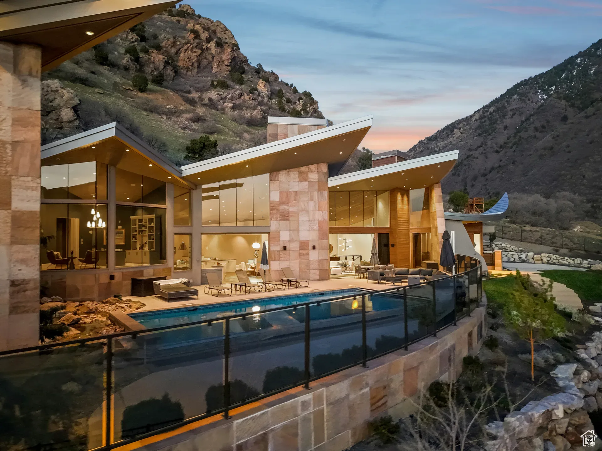 A luxury mountain home's infinity pool and patio glowing at dusk with the hillside behind it