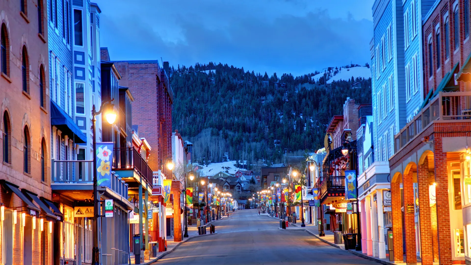 Park City's Old Town Main Street at twilight, brick storefronts and lit street lamps beneath snow-capped peaks