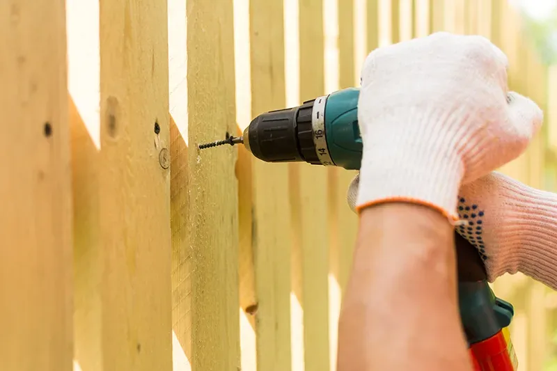 Fence repair by a handyman in San Antonio: a gloved hand using a power drill to secure a panel on a wood privacy fence in bright daylight