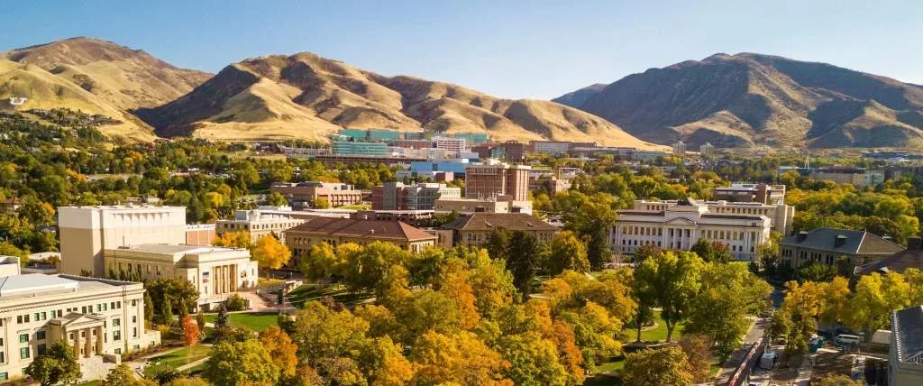 A high autumn aerial over the Salt Lake Valley, the university foothills and tree canopy beneath golden peaks