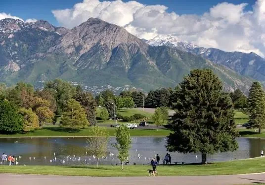 Sugar House Park on a sunny day, a lake in the foreground with mature trees and snow-capped mountains behind