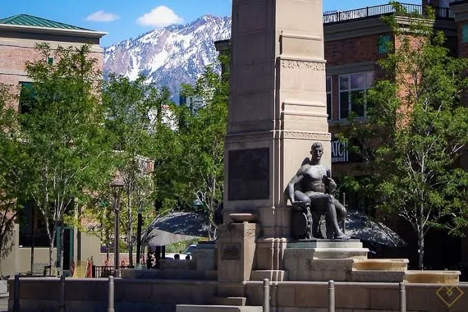 A stone monument with a bronze sculpture in a Sugar House plaza, brick storefronts and green trees around it