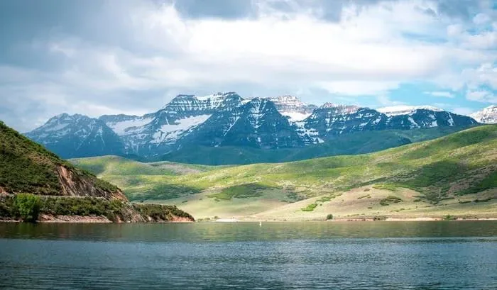 A wide view over an alpine lake in the Heber Valley, snow-capped mountains and green rolling hills beyond the calm water, partly cloudy daylight